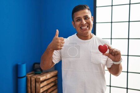 Photo for Hispanic young man holding red heart at gym smiling happy and positive, thumb up doing excellent and approval sign - Royalty Free Image