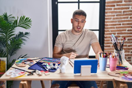 Photo for Young hispanic painter man doing online call with tablet scared and amazed with open mouth for surprise, disbelief face - Royalty Free Image