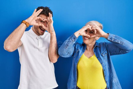 Photo for Young brazilian mother and son standing over blue background doing heart shape with hand and fingers smiling looking through sign - Royalty Free Image
