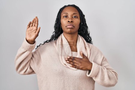 Foto de Mujer africana de pie sobre fondo blanco jurando con la mano en el pecho y la palma abierta, haciendo una promesa de lealtad juramento - Imagen libre de derechos