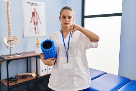 Photo for Young physiotherapist woman holding foam roll at the clinic with angry face, negative sign showing dislike with thumbs down, rejection concept - Royalty Free Image