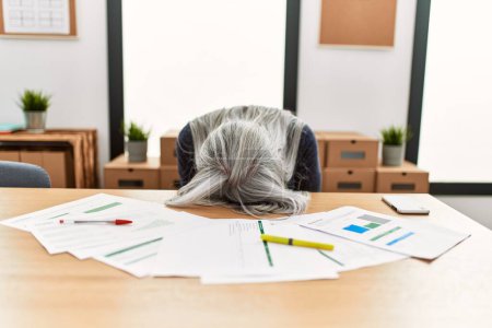 Foto de Mujer de pelo gris de mediana edad trabajadora de negocios estresada con la cabeza en la mesa en la oficina - Imagen libre de derechos