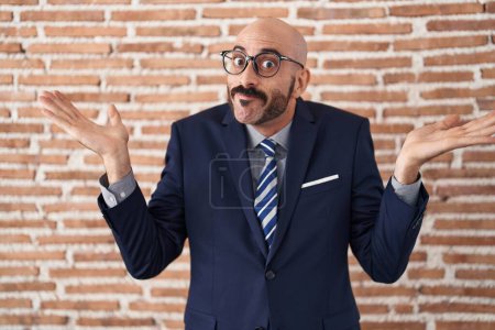 Photo for Bald man with beard wearing business clothes and glasses clueless and confused expression with arms and hands raised. doubt concept. - Royalty Free Image