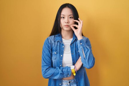 Téléchargez les photos : Jeune femme asiatique debout sur fond jaune bouche et lèvres fermées comme zip avec les doigts. secret et silencieux, tabou parlant - en image libre de droit
