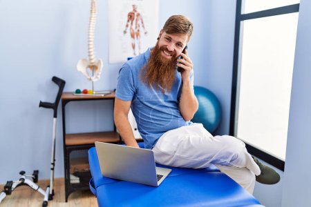 Photo for Young redhead man wearing physiotherapist uniform using laptop and talking on the smartphone at physiotherapy clinic - Royalty Free Image