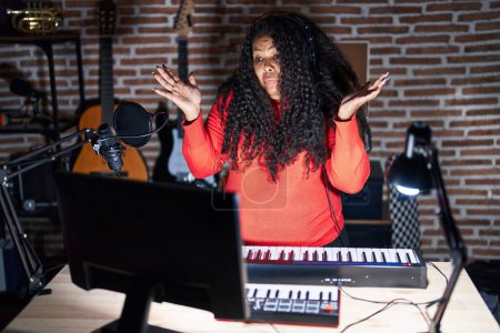 Plus size hispanic woman playing piano at music studio clueless and confused expression with arms and hands raised. doubt concept.