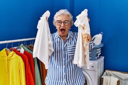 Photo for Senior woman with grey hair holding clean white t shirt and t shirt with dirty stain smiling and laughing hard out loud because funny crazy joke. - Royalty Free Image
