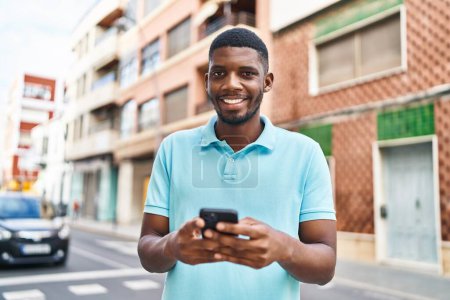 Foto de Joven afroamericano hombre sonriendo confiado usando teléfono inteligente en la calle - Imagen libre de derechos
