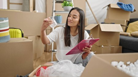 Photo for Young beautiful hispanic woman unpacking cardboard box checking on checklist at new home - Royalty Free Image