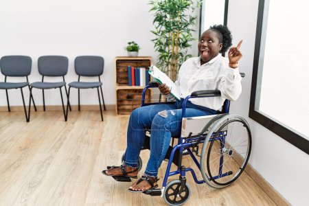 Téléchargez les photos : Jeune femme noire assise en fauteuil roulant dans la salle d'attente souriant étonné et surpris et pointant vers le haut avec les doigts et les bras levés. - en image libre de droit