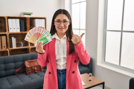 Photo for Young latin woman working at consultation office holding money pointing thumb up to the side smiling happy with open mouth - Royalty Free Image