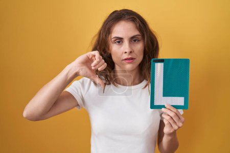Photo for Beautiful brunette woman holding l sign for new driver with angry face, negative sign showing dislike with thumbs down, rejection concept - Royalty Free Image