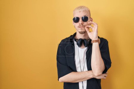 Téléchargez les photos : Jeune homme caucasien portant des lunettes de soleil debout sur fond jaune bouche et lèvres fermées comme zip avec les doigts. secret et silencieux, tabou parlant - en image libre de droit