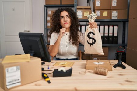 Photo for Young hispanic woman working at small business ecommerce holding money bag serious face thinking about question with hand on chin, thoughtful about confusing idea - Royalty Free Image