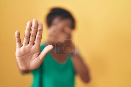 Téléchargez les photos : Femme africaine avec dreadlocks debout sur fond jaune couvrant les yeux avec les mains et faire arrêter geste avec expression triste et peur. concept embarrassé et négatif. - en image libre de droit