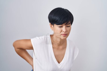 Foto de Joven mujer asiática con pelo corto de pie sobre fondo aislado sufriendo de dolor de espalda, tocando la espalda con la mano, dolor muscular - Imagen libre de derechos