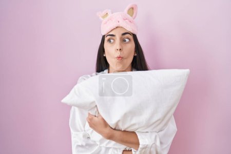 Photo for Young brunette woman wearing sleep mask and pajama hugging pillow making fish face with mouth and squinting eyes, crazy and comical. - Royalty Free Image