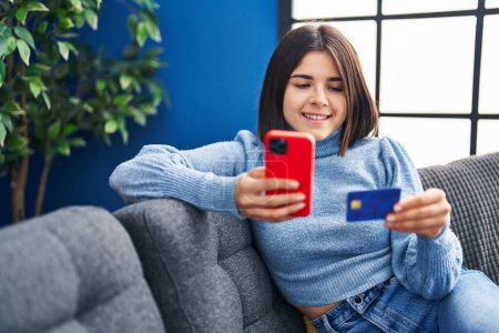Foto de Joven mujer hispana hermosa usando teléfono inteligente y tarjeta de crédito sentado en el sofá en casa - Imagen libre de derechos