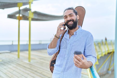 Téléchargez les photos : Jeune musicien hispanique parlant sur smartphone boire du café au bord de la mer - en image libre de droit