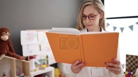 Photo for Young blonde woman preschool teacher smiling confident reading book at kindergarten - Royalty Free Image