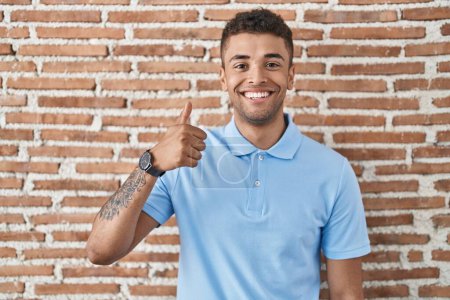 Téléchargez les photos : Jeune homme brésilien debout sur un mur de briques faisant des pouces heureux geste avec la main. approuvant l'expression regardant la caméra montrant le succès. - en image libre de droit