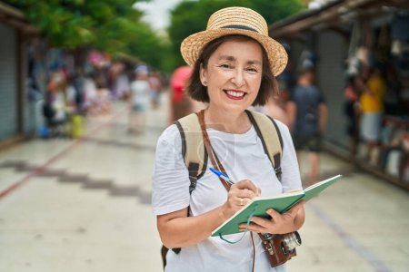 Foto de Mujer de mediana edad turista sonriendo escritura segura en el cuaderno en el mercado callejero - Imagen libre de derechos