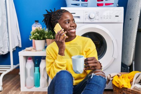 Photo for African american woman talking on the smartphone drinking coffee waiting for washing machine at laundry room - Royalty Free Image