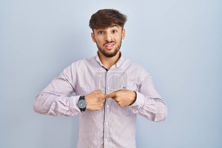 Téléchargez les photos : Homme arabe avec la barbe debout sur fond bleu à la hâte pointant pour regarder le temps, l'impatience, bouleversé et en colère pour retard de délai - en image libre de droit