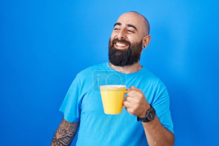 Foto de Joven hombre hispano con barba y tatuajes bebiendo una taza de café mirando hacia un lado con sonrisa en la cara, expresión natural. riendo confiado. - Imagen libre de derechos