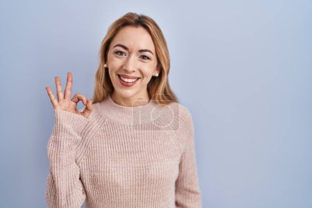 Foto de Mujer hispana de pie sobre fondo azul sonriendo positiva haciendo signo ok con la mano y los dedos. expresión exitosa. - Imagen libre de derechos