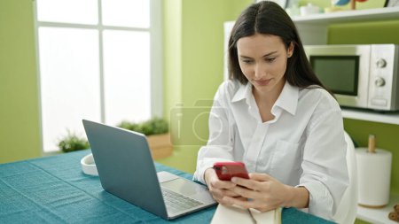 Téléchargez les photos : Jeune belle femme hispanique utilisant smartphone et ordinateur portable assis sur la table à manger - en image libre de droit