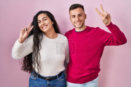 Foto de Joven pareja hispana de pie sobre fondo rosa sonriendo mirando a la cámara mostrando los dedos haciendo señal de victoria. número dos. - Imagen libre de derechos
