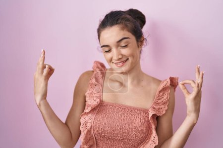 Téléchargez les photos : Jeune adolescente debout sur fond rose se détendre et sourire les yeux fermés en faisant geste de méditation avec les doigts. concept de yoga. - en image libre de droit