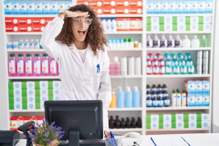 Photo for Hispanic woman with curly hair working at pharmacy drugstore very happy and smiling looking far away with hand over head. searching concept. - Royalty Free Image