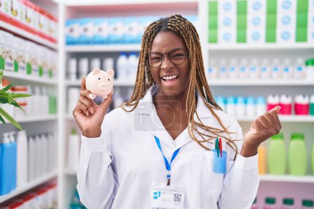 Téléchargez les photos : Femme afro-américaine avec des cheveux tressés travaillant à la pharmacie pharmacie tenant tirelire criant fier, célébrant la victoire et le succès très excité avec le bras levé - en image libre de droit