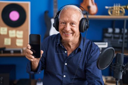 Téléchargez les photos : Homme âgé avec les cheveux gris montrant écran de smartphone au studio de musique regardant positif et heureux debout et souriant avec un sourire confiant montrant les dents - en image libre de droit