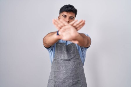 Téléchargez les photos : Jeune homme hispanique portant tablier sur fond blanc expression de rejet traversant les bras et les paumes faisant signe négatif, visage en colère - en image libre de droit