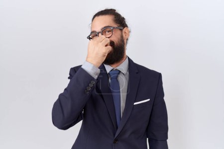 Foto de Hombre hispano con barba vistiendo traje y corbata oliendo algo apestoso y asqueroso, olor intolerable, conteniendo la respiración con los dedos en la nariz. mal olor - Imagen libre de derechos