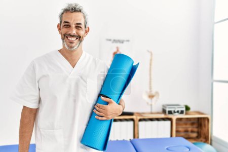 Photo for Middle age grey-haired man physiotherapist smiling confident holding yoga mat at rehab clinic - Royalty Free Image