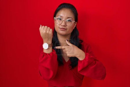 Téléchargez les photos : Asiatique jeune femme debout sur fond rouge à la hâte pointant pour regarder le temps, impatience, regardant la caméra avec une expression détendue - en image libre de droit