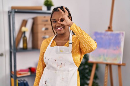 Foto de Mujer afroamericana con trenzas en el estudio de arte sonriendo feliz haciendo bien signo con la mano en el ojo mirando a través de los dedos - Imagen libre de derechos