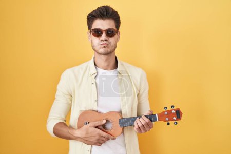 Photo for Young hispanic man playing ukulele over yellow background puffing cheeks with funny face. mouth inflated with air, catching air. - Royalty Free Image