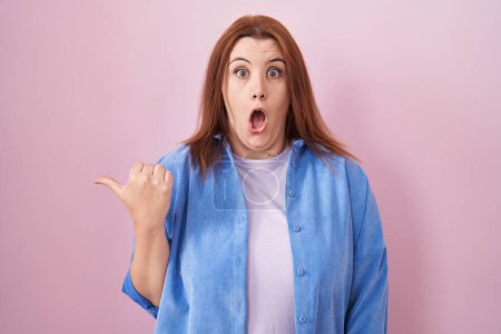 Photo for Young hispanic woman with red hair standing over pink background surprised pointing with hand finger to the side, open mouth amazed expression. - Royalty Free Image