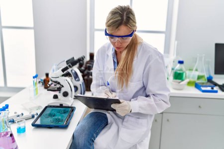 Photo for Young blonde woman wearing scientist uniform writing on clipboard looking embryion images at laboratory - Royalty Free Image