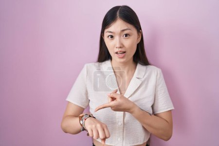 Téléchargez les photos : Chinois jeune femme debout sur fond rose à la hâte pointant pour regarder le temps, impatience, bouleversé et en colère pour retard de délai - en image libre de droit