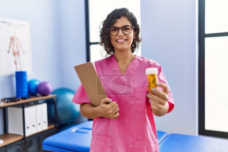 Téléchargez les photos : Young latin woman wearing physiotherapist uniform holding pills at clinic - en image libre de droit