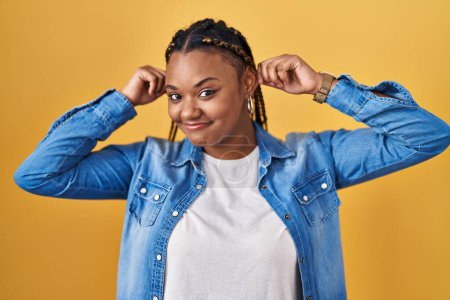 Téléchargez les photos : Femme afro-américaine avec des tresses debout sur fond jaune souriant tirant les oreilles avec les doigts, geste drôle. problème d'audition - en image libre de droit