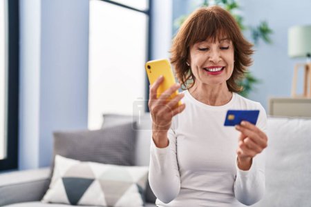 Foto de Mujer de mediana edad con teléfono inteligente y tarjeta de crédito sentado en el sofá en casa - Imagen libre de derechos