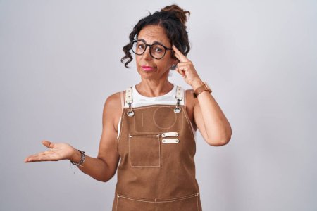 Photo for Middle age woman wearing apron over white background confused and annoyed with open palm showing copy space and pointing finger to forehead. think about it. - Royalty Free Image