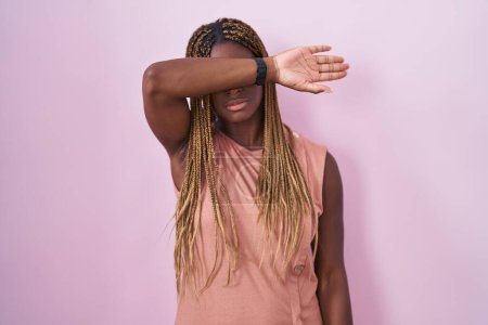 Foto de Mujer afroamericana con cabello trenzado de pie sobre fondo rosa cubriendo los ojos con el brazo, luciendo seria y triste. concepto de ocultamiento y rechazo sin visión - Imagen libre de derechos